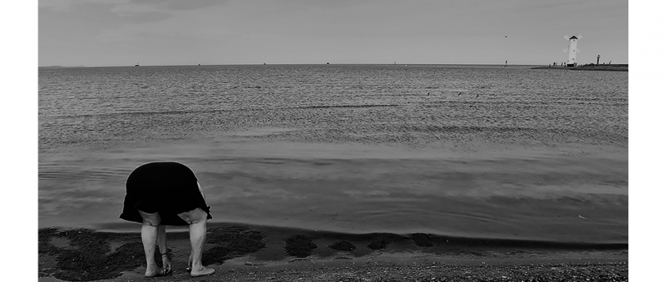 Fotografia Anny Lewańskiej pokazująca samotną, starszą kobietę nad brzegiem morza. Pochylona postać w akcie zbierania prawdopodobnie muszelek.