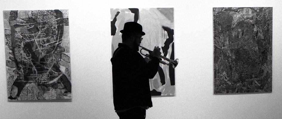 zdjęcie Artura Tuźnika podczas próby. Gra na trąbce w galerii, zdjęcie czarno-białe na tle obrazów abstrakcyjnych Gabi Buzek 