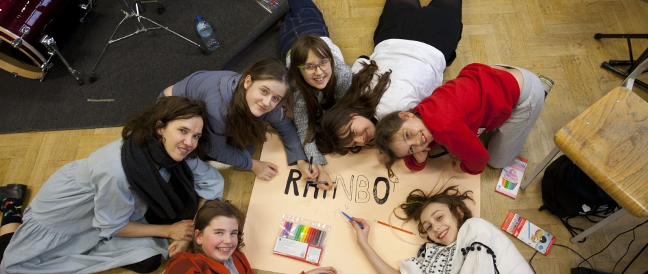 zdjęcie dziewczynek uczestniczących w warsztatach z tutorką Olą Rzepką. Wspólnie tworzą plakat zespołu. Śmieją się. Widać fragment napisu Rainbo... na plakacie